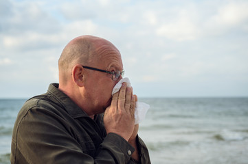 A man stands by the sea and uses a handkerchief, he has an allergy or severe flu. Concept: Runny nose or protection against infections
