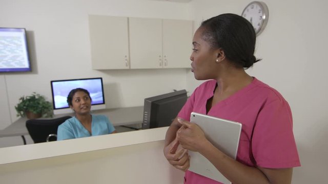 Two female nurses talking at hospital nurses station, one holding a tablet