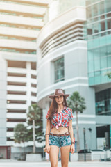 Portrait of young woman in checkered shirt, denim shorts and cowboy hat. Country girl came to a big city from a small village. Vertical shot. Front view. Modern buildings in the background