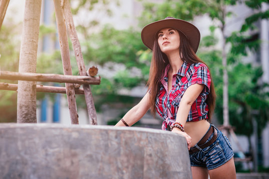 Portrait of caucasian young woman in checkered shirt, denim shorts and cowboy hat. She is dreaming about life in a big city while standing outdoors in a countryside. Horizontal shot. Selective focus