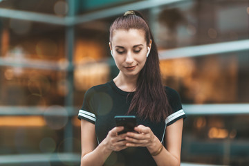 Portrait of young woman looking at the screen of her phone with a smile while standing outdoors and...