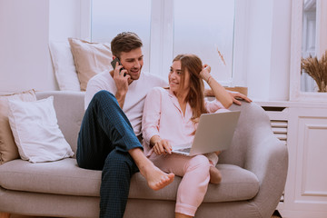 Couple in the morning making a order from the phone and watching on the notebook they are happy sitting on the sofa in pajamas