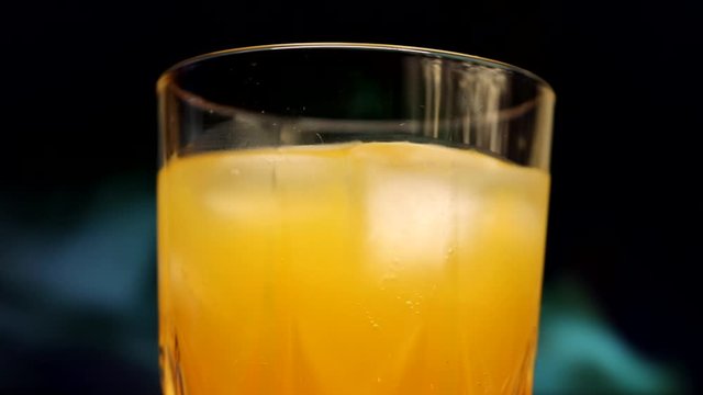 Slowly rotating glass with yellow-orange sweet multifruit juice and ice cubes. Glass with juice on a background of colored smoke.