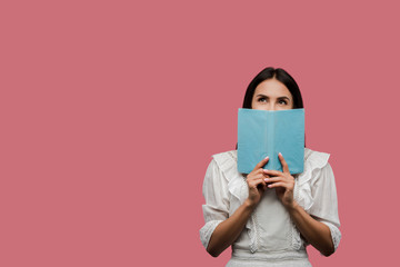 young woman in dress covering face with book isolated on pink