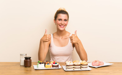 Obraz na płótnie Canvas Young woman with lots of different mini cakes in a table giving a thumbs up gesture