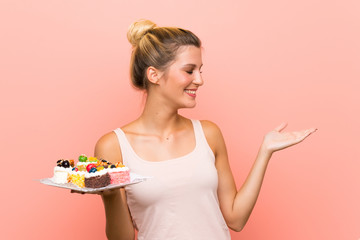 Young blonde woman holding lots of different mini cakes with surprise facial expression