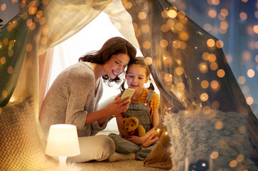 family, hygge and technology concept - happy mother and little daughter with smartphone in kids tent at night at home