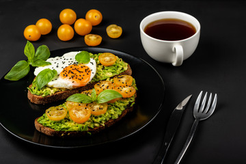 Open sandwiches with avocado guacamole, yellow cherry tomatoes, fried egg and basil on a black plate on the black background. Healthy food or snack