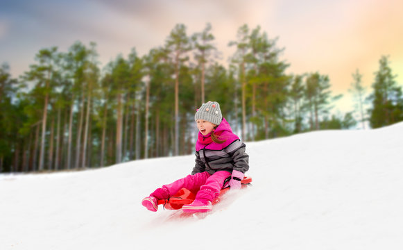 childhood, sledging and season concept - happy little girl sliding down on sled outdoors over winter forest background
