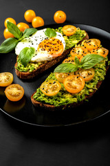 Open sandwiches with avocado guacamole, yellow cherry tomatoes, fried egg and basil on a black plate on the black background. Healthy food or snack
