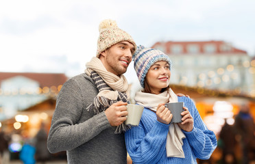 Obraz premium people and winter holidays concept - happy romantic couple in knitted hats and scarves with mugs over christmas market in old town of tallinn city background