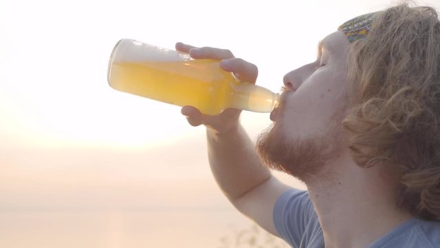 Portrait of hipster young guy drinking beer or limonade on the background of awesome sunset on the beach during picnic