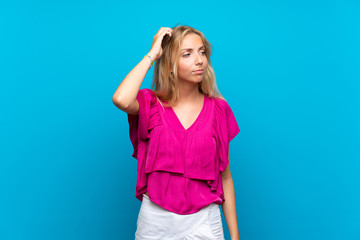 Blonde young woman over isolated blue background having doubts while scratching head