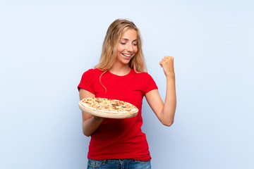 Young blonde woman holding a pizza over isolated blue wall celebrating a victory