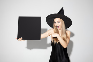 Young blonde woman in black hat and costume on white background. Attractive caucasian female model. Halloween, black friday, cyber monday, sales, autumn concept. Copyspace. Holding black copyspace.