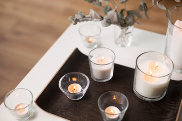 decoration, hygge and cosiness concept - burning white fragrance candles on tray and branches of eucalyptus populus on table