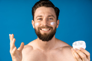 Man applying moisturizer cream on his face skin on blue background. Super satisfied.
