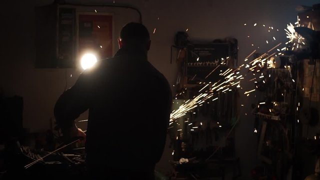A man sprays iron with a circular saw in his garage. A man crafts his own in a small garage. Sparks fly everywhere