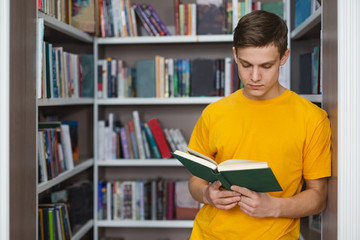 Smart male student reading book between bookshelves