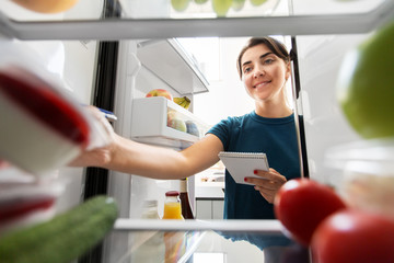 healthy eating and diet concept - woman opening fridge and making list of necessary food at home...