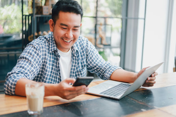 Asian young man in blue shirt working with laptop computer and mobile phone with online job or shopping online happy and smile face in coffee shop cafe