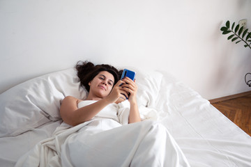 woman with smartphone in bed with white sheets