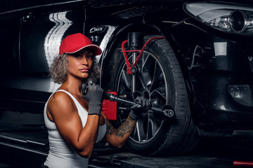 Naklejka na ściany i meble Blond strong woman is doing man's job, fixing car's wheel with wrench at auto service.