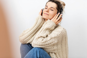 Image of European young woman in beige loose sweater, relaxing with closed eyes, listening to favourite songs via earphones, using music app. Pretty female enjoying different audio tracks on headphone