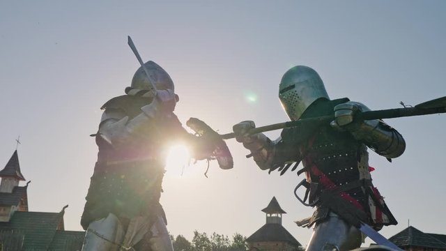 Battle of two knights in armor near wooden fortress. Knights are fighting against blue sky with sun.