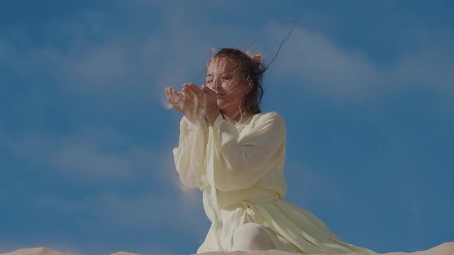 Woman in light yellow dress on sand dune in fornt of blue sky pouring sand from hands on wind. Slow motion