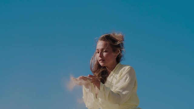 Woman in light yellow dress on sand dune in fornt of blue sky pouring sand from hands on wind. Slow motion