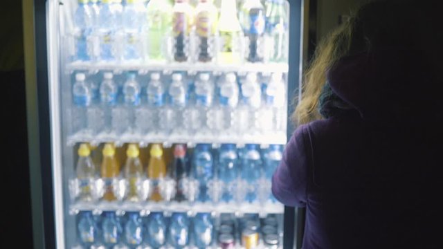 View from the back of young woman in purple coat choosing a drink from vending machine. Stock footage. Beverage vending machine selling water in bottles
