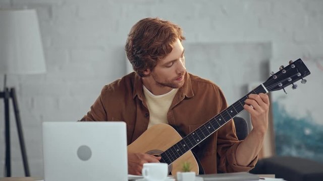 redhead man playing acoustic guitar while looking at laptop screen