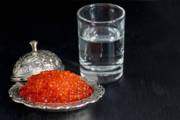 Red caviar in silver bowl with Russian vodka on black background