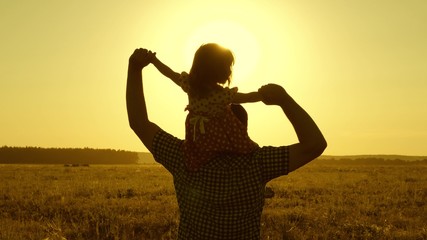 Dad carries beloved baby on his shoulders walking around field at sunset. little daughter riding with dad on his shoulders in park. child with parent walks in evening at sunset of sun. Slow motion.