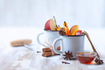wo white metal mugs with ingredients for hot mulled wine - traditional christmas beverage. Ripe apple and orange, spices and honey. Corkscrew to open wine bottle. Holiday festive mood, rustic style