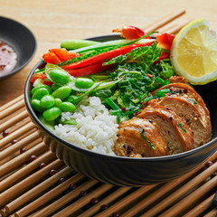Buddha bowl meal with teriyaki chicken fillet, fresh vegetable salad, stew cabbage, edamame and basmati rice on a wooden table. Delicious dish in a black bowl.
