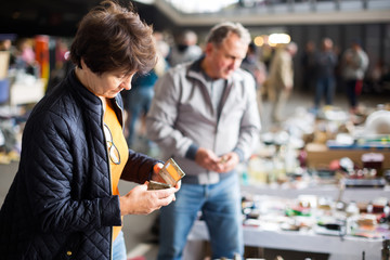 Positive elderly woman choosing interesting souvenirs at flea market