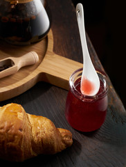A spoon in a jar of raspberry jam and a croissant on a dark background Board. The jam drips from the spoon. Rustic style