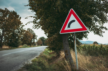 Image - Illuminated European traffic sign (red triangle) -curved road, green meadow and field on background on sunset. Beware of curve.