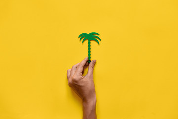 Hand holding palm tree plastic toy. Yellow background. Summer travel. Ecology idea. Copy space
