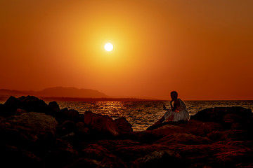 Fototapeta na wymiar Silhouette of a young woman sitting on a stone beach and using a smartphone on a beautiful sea sunset background