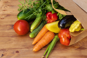 Green, yellow and red vegetables in a paper bag lying on their side on a brown wooden board, side view from above