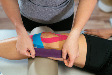 Photo detail of the hands of a physiotherapist woman gluing purple medical tape on another celestial tape and another pink one on the knee of a patient. Concept of muscle health and relaxation. - 288966163