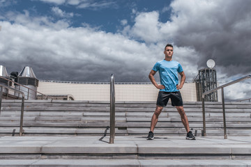 Athletic male athlete, summer in city stands on stairs, in sportswear, warm-up workout fitness in morning, free space for motivation text. Active lifestyle of young people, powerful and stylish man.