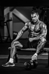 Fototapeta na wymiar Bodybuilder sitting on Bench at the Gym With One Lef Elevated on a Dumbbell