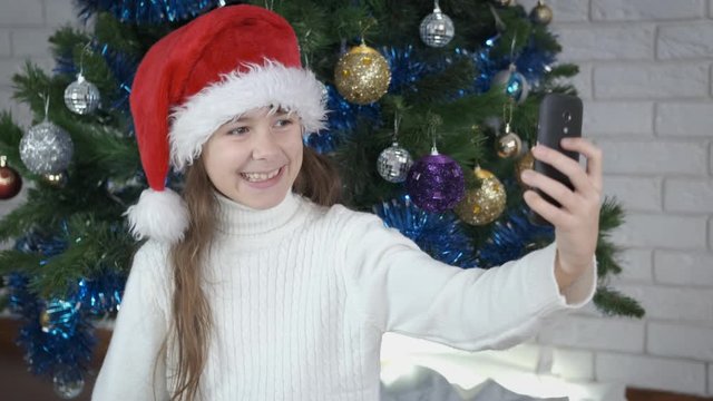 Girl grimaces for photo. Happy little girl is photographed on the phone against the background of a Christmas tree.