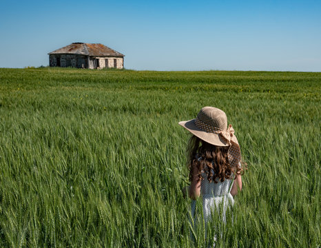 Girl in straw hat standing in green wheat field looking at stone schoolhouse in the distance