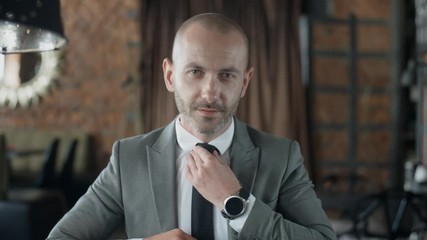 Close up portrait of businessman in business suit straightens tie, handsome stylish man sitting at table in loft design cafe with brick walls. He looks at camera and smiles, day light