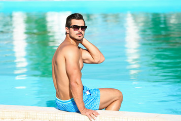 Handsome young man sitting at swimming pool edge on sunny day